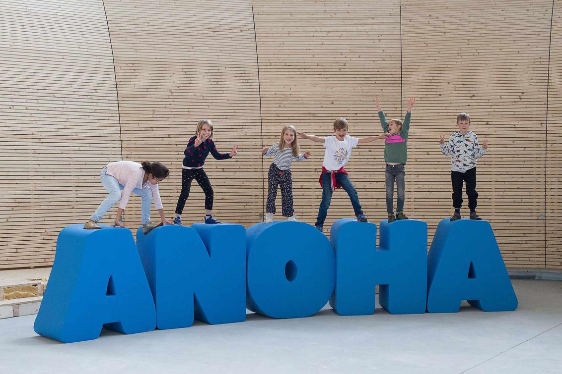 Kinder turnen auf lebensgroßen Buchstaben, die den Schriftzug ANOHA bilden