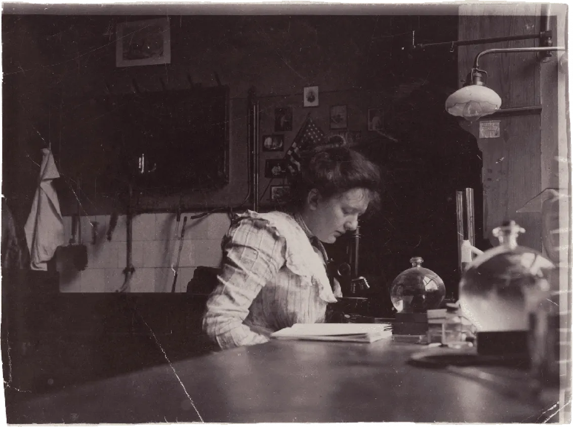 Historical black and white photograph of a young woman reading bent over a table.