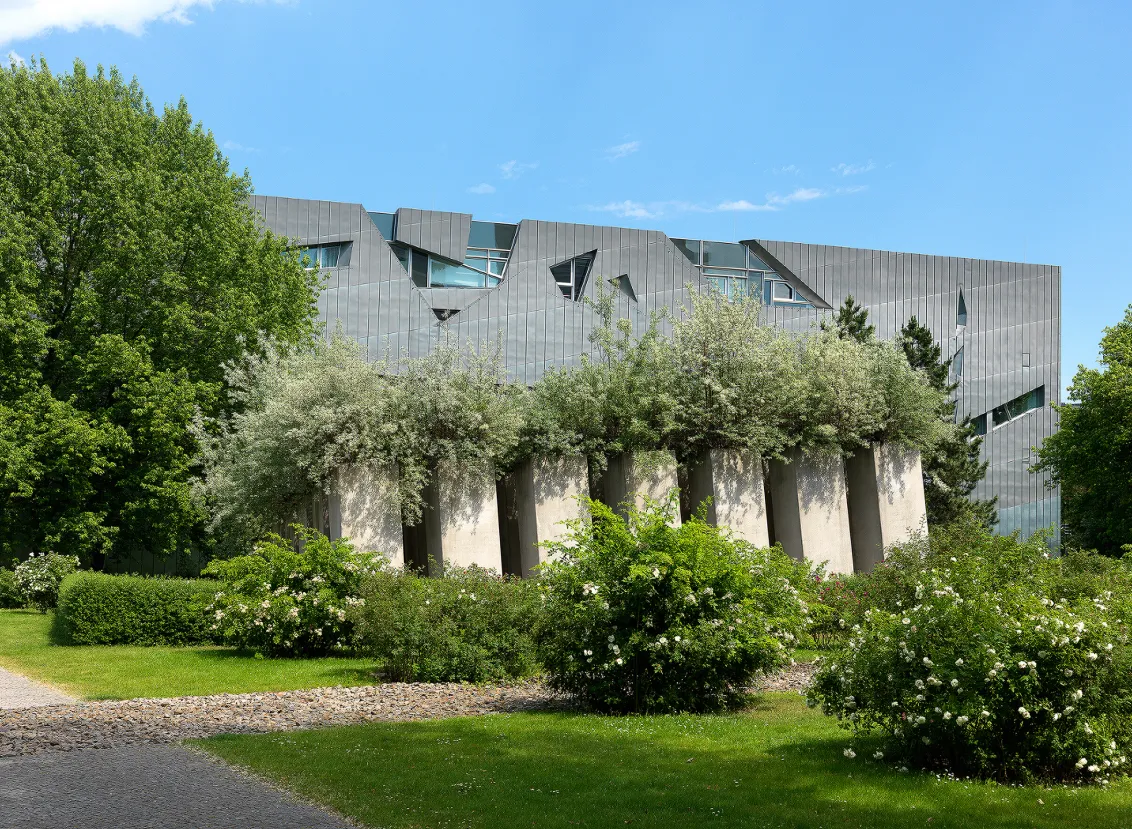 Rosenhain vor dem Garten des Exil mit quadratisch angeordneten Stelen auf einer schiefen Ebene, daraus wachsen Ölweiden