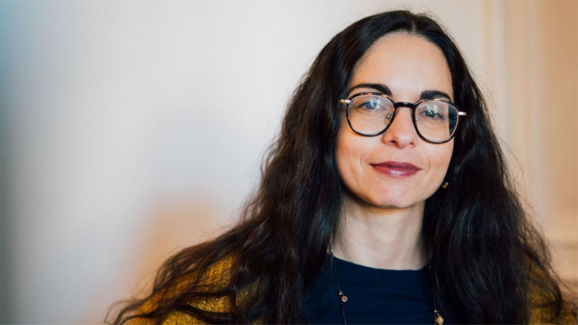 A woman with dark, long hair and glasses looks friendly into the camera.