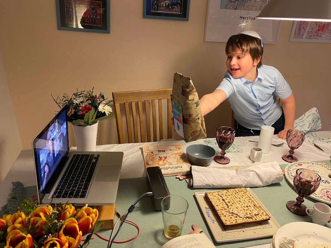 A boy with kippah holds a brown paper bag into the camera from a laptop.