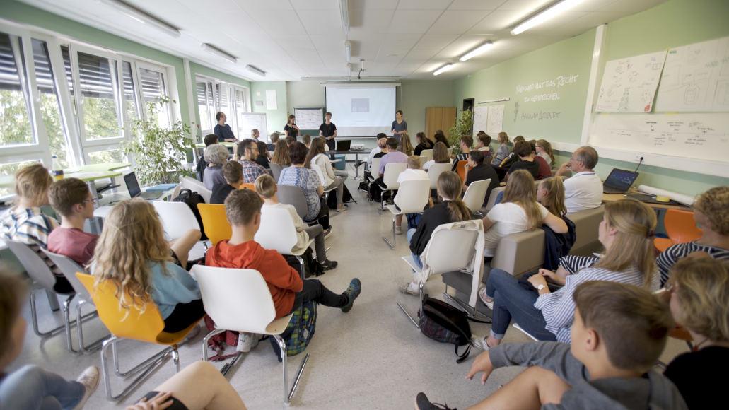Young people and adults sit on rows of chairs in a large room listening to three young people who are speaking in front of a projector screen.