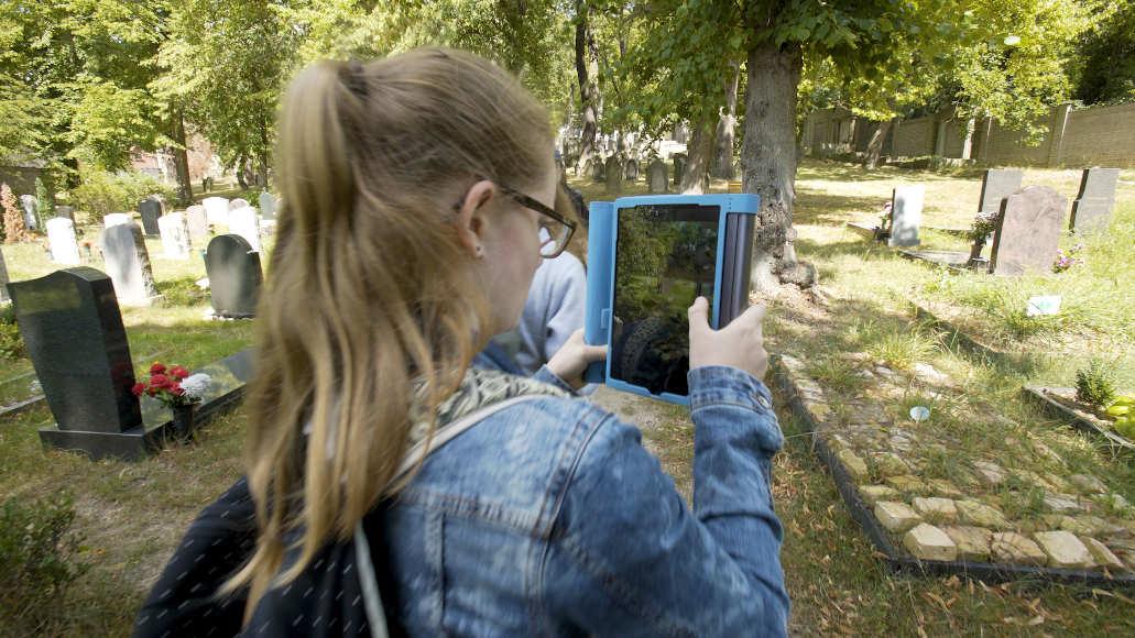 A girl uses a tablet to take a photograph of a tombstone in a cemetery.