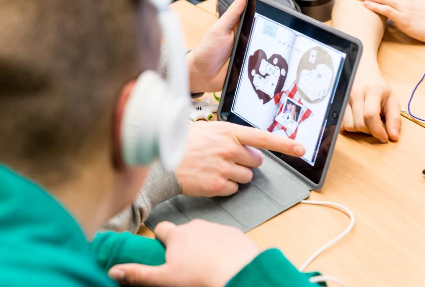 Ein Hand wischt über das iPad während ein Schüler mit Kopfhörern auf das Display sieht.