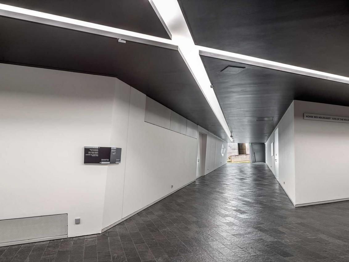 Long corridor with white walls and black floor, black ceilings, lights forming a cross.
