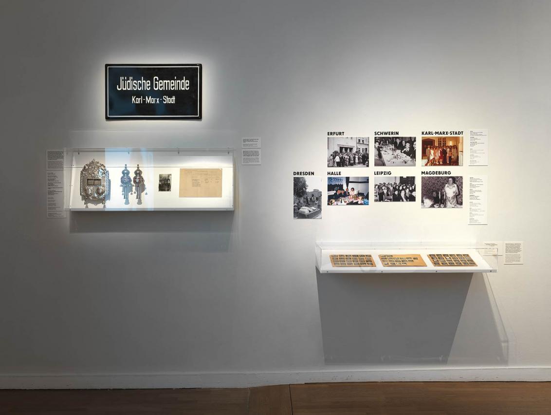 View of an exhibition wall with a plaque of the Jewish Community Karl-Marx-Stadt, ceremonial objects in a showcase and photos of other Jewish communities in the GDR.