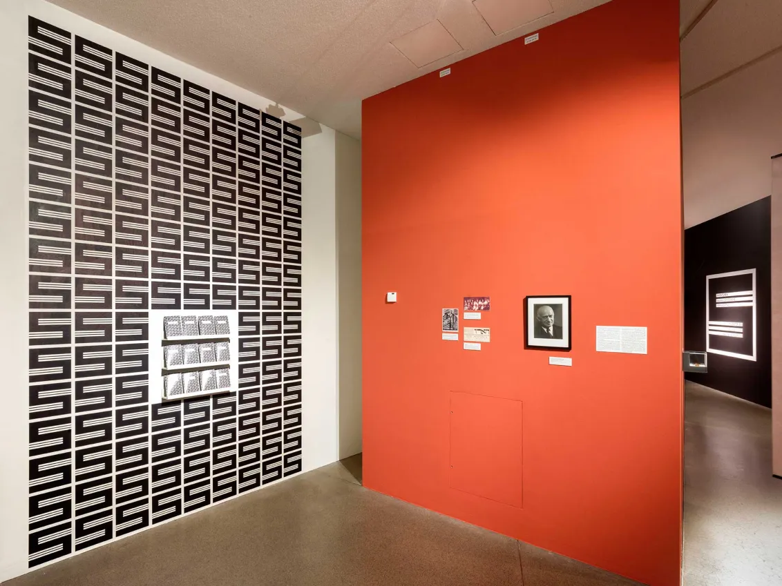 View into an exhibition room, with a central orange-colored wall on which hangs a framed portrait photo of Salman Schocken, on the wall to the left the S-shaped Schocken logo in black and white several times as well as some brochures.
