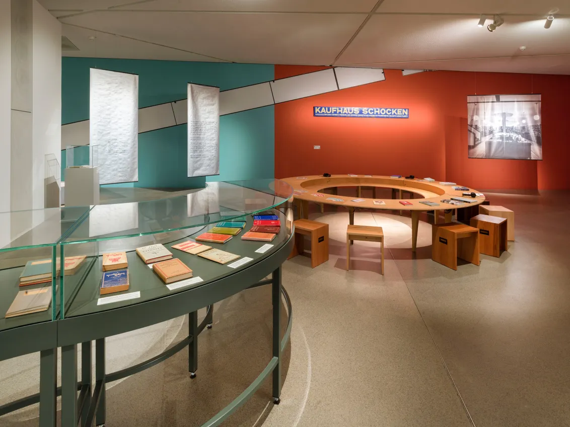 View into an exhibition room, in the foreground a round green display case with books, in the background a round wooden table with chairs, behind it an orange-painted wall with a sign for the Schocken department store.