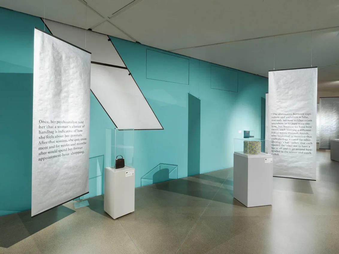 View into an exhibition room with a light blue wall, a handbag and a hatbox are displayed on white pedestals, texts are written on banners hanging in the room.