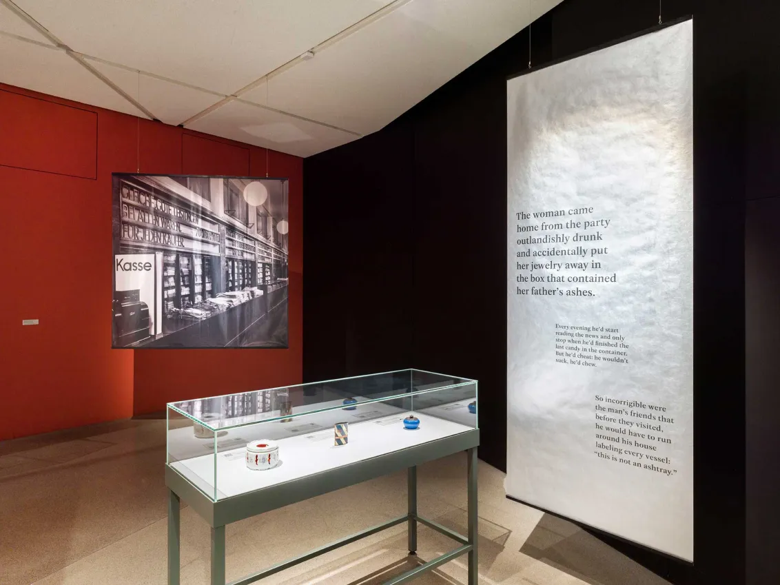View into an exhibition room with a display case showing a teapot, a tin can and a porcelain can, with a close-up of a department store in the background.