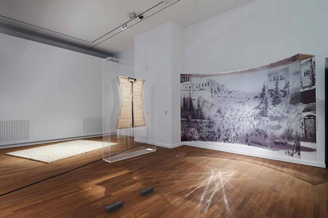 In front of one wall hangs a curved installation made from a black and white photograph, next to it is a display case containing a work of art that looks like a sleeveless top. In the background, a white surface can be seen on the floor.