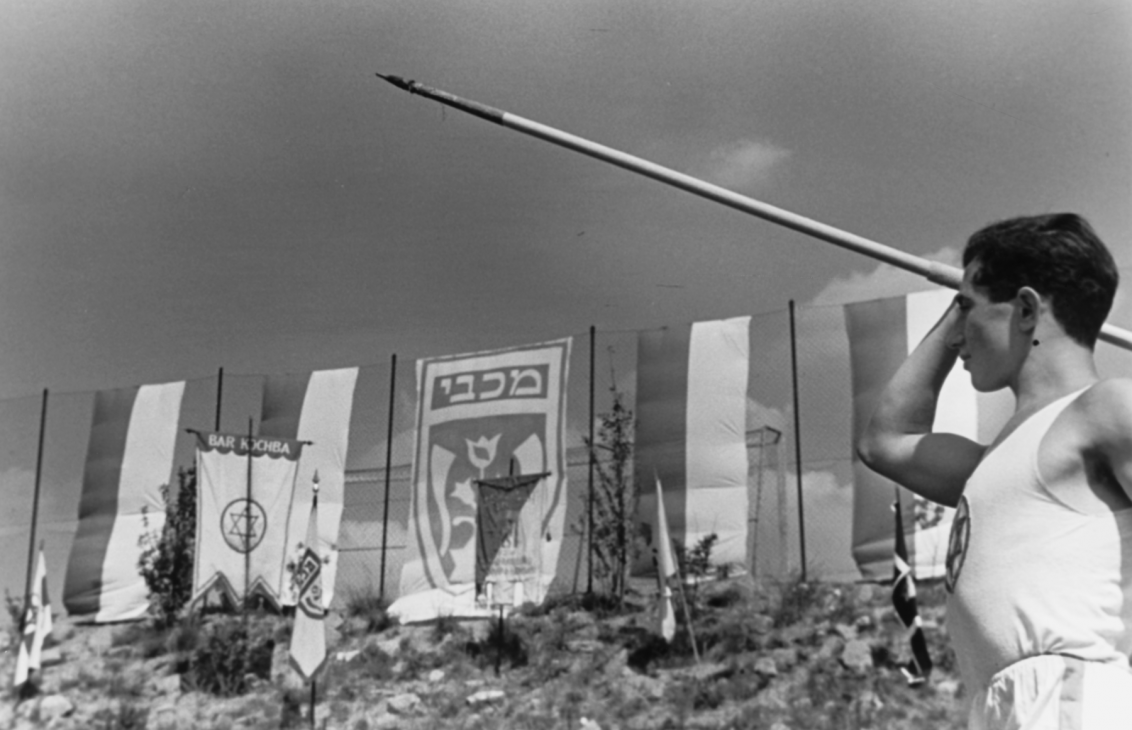  Young man holding a spear in a throwing position above his head. In the background flags.