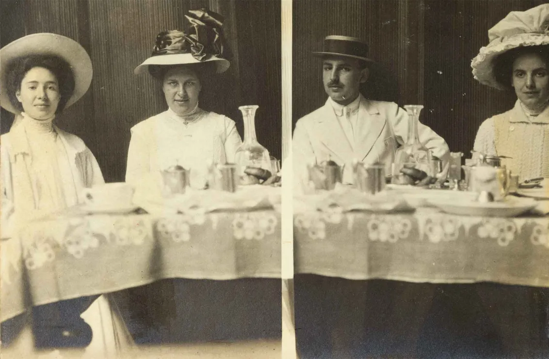 Schwarz-Weiß-Foto von vier jungen Personen an einer gedeckten Tafel. Alle tragen Hüte und blicken kess in die Kamera