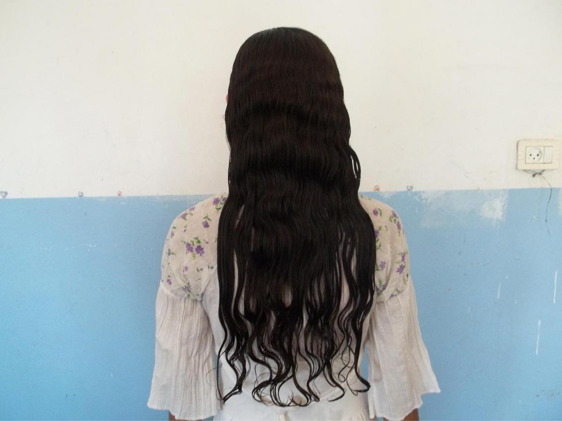 Photo of a woman from behind, from head to hip, with long dark hair in front of a blue-white wall