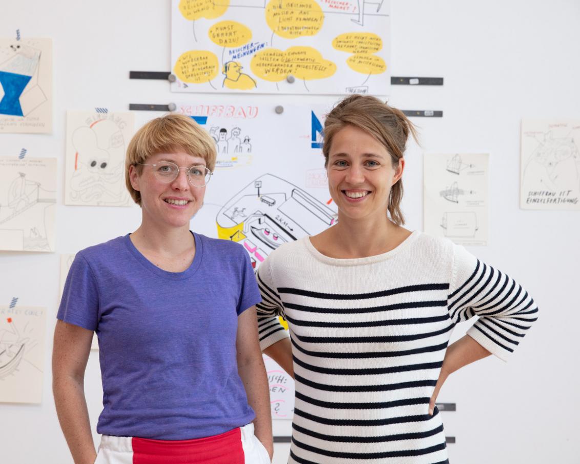 Two women with blond hair stand in front of a wall with post-its and smile.