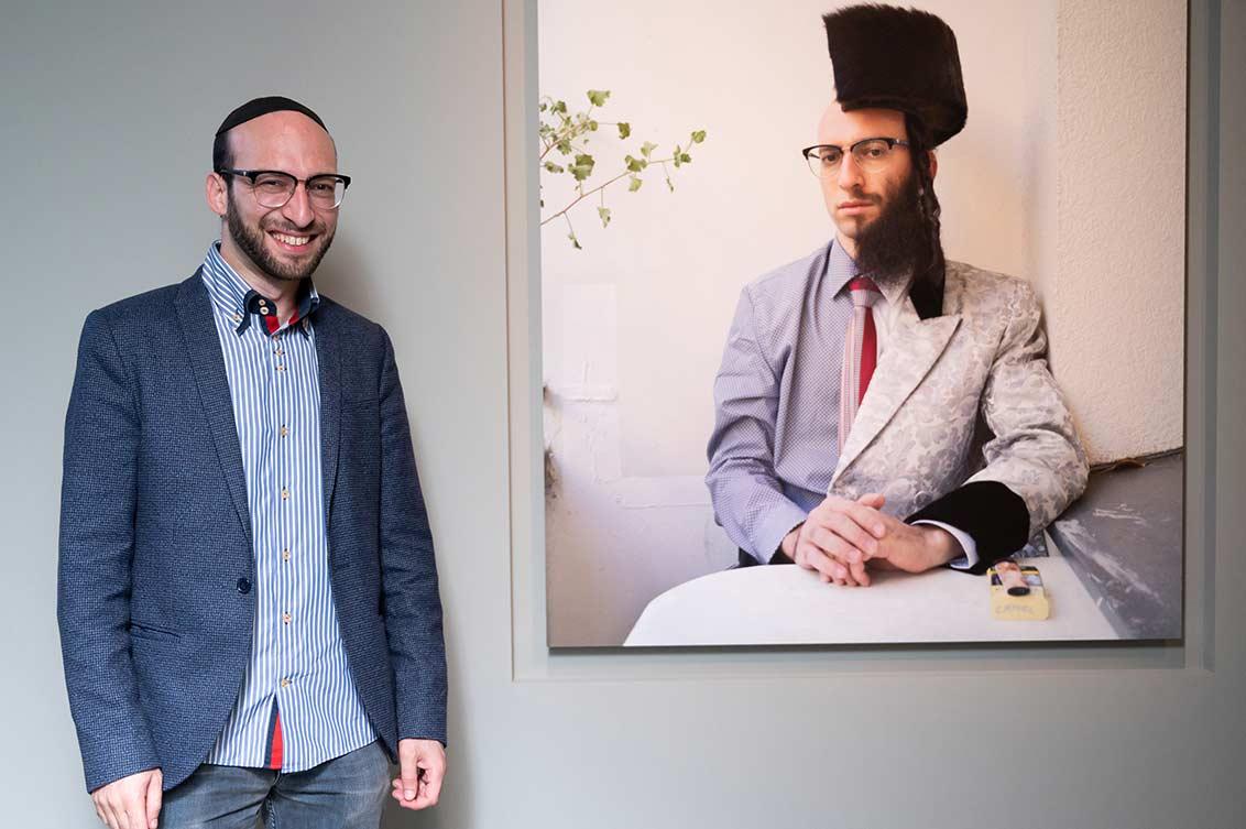 A man in a suit with a kippah stands next to a portrait of himself on display; on the portrait on the wall he is cut in half: one half wears a beard and shtreimel, one half is shaved and bald.