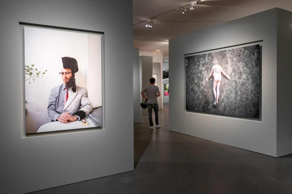 A visitor walks between two walls of large-scale portraits: one shows a man cut in two, as it were, with half a beard and shtreimel, one shows a naked man lying prone on the floor