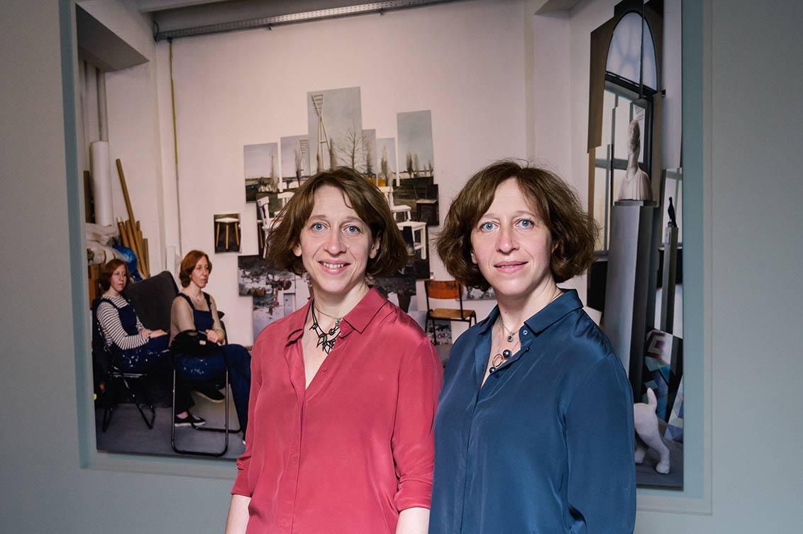 Twin sisters in blue and red blouse in the exhibition ZERHEILT in front of a portrait of them in their studio