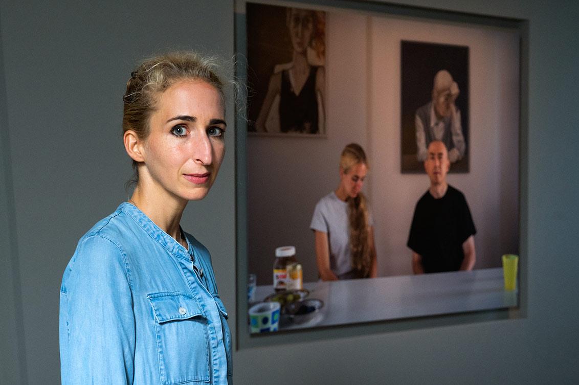 A blonde woman in a denim dress stands in the exhibition next to a photograph of herself and her partner sitting at the breakfast table, in the background two paintings on the wall
