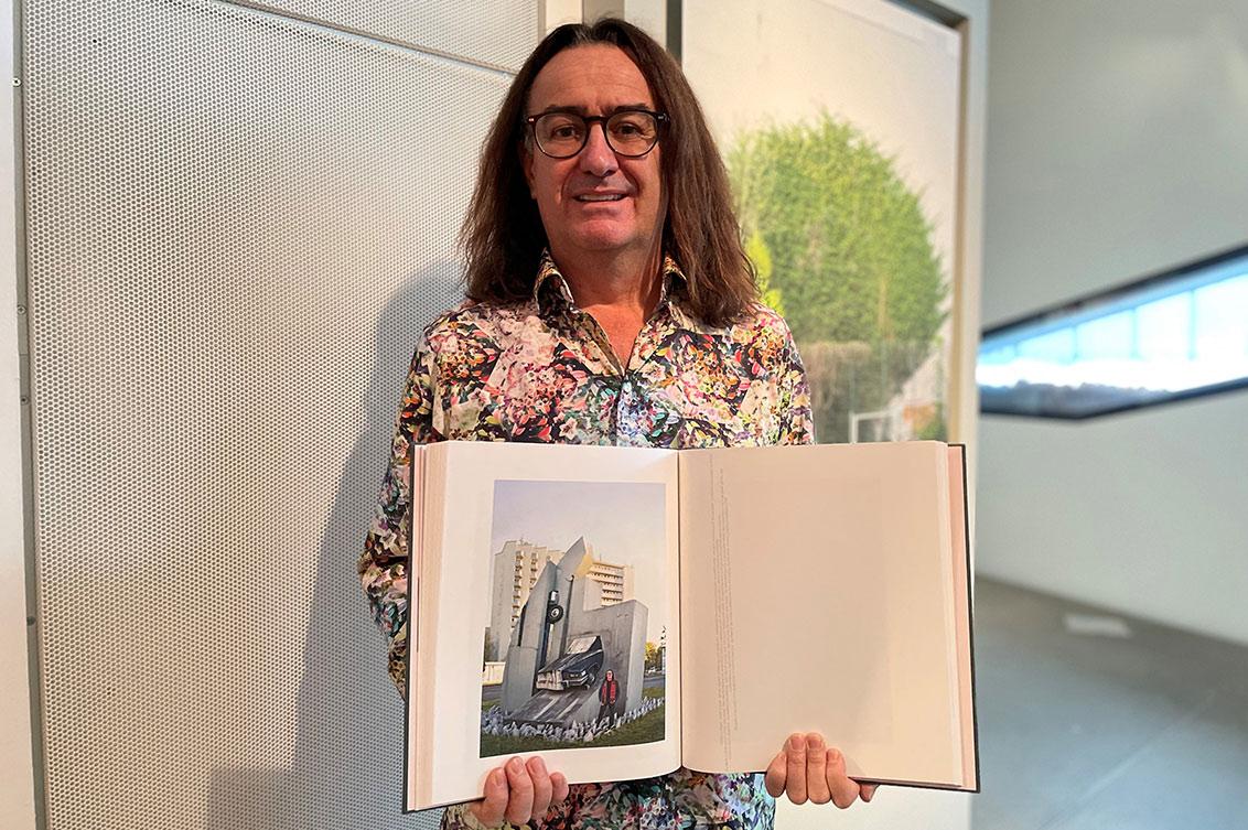 A man with long hair, glasses and colorful patterned shirt stands in an exhibition and holds an open book into the camera, in which is a portrait photo of him