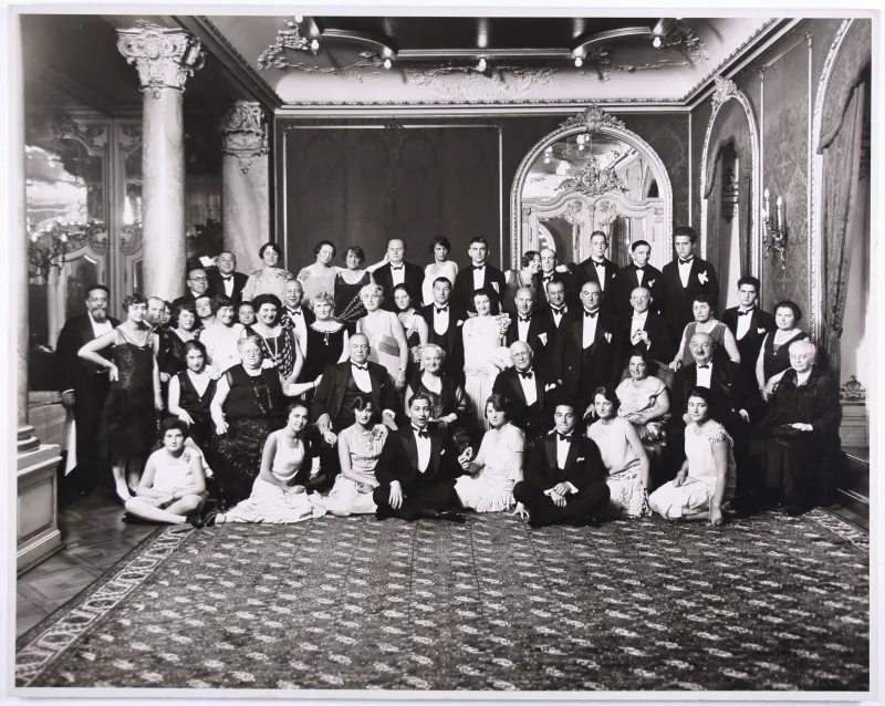 Black-and-white photo. Large group photo of a festive gathering. The ladies and gentlemen are wearing elegant clothing and are standing in a large room.