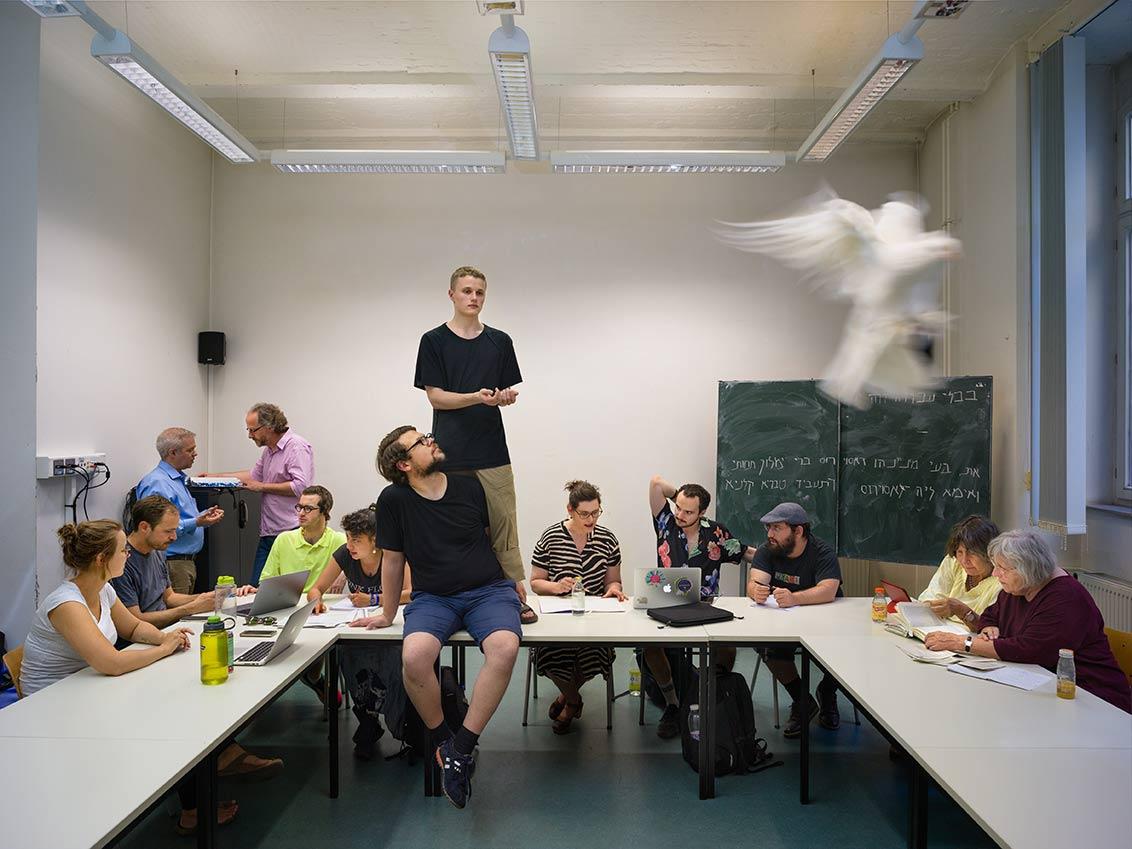 Schülerinnen und Schüler einer Talmud-Klasse schauen auf Texte oder sich gegenseitig an, während im Vordergrund des Bilds eine weiße Taube fliegt