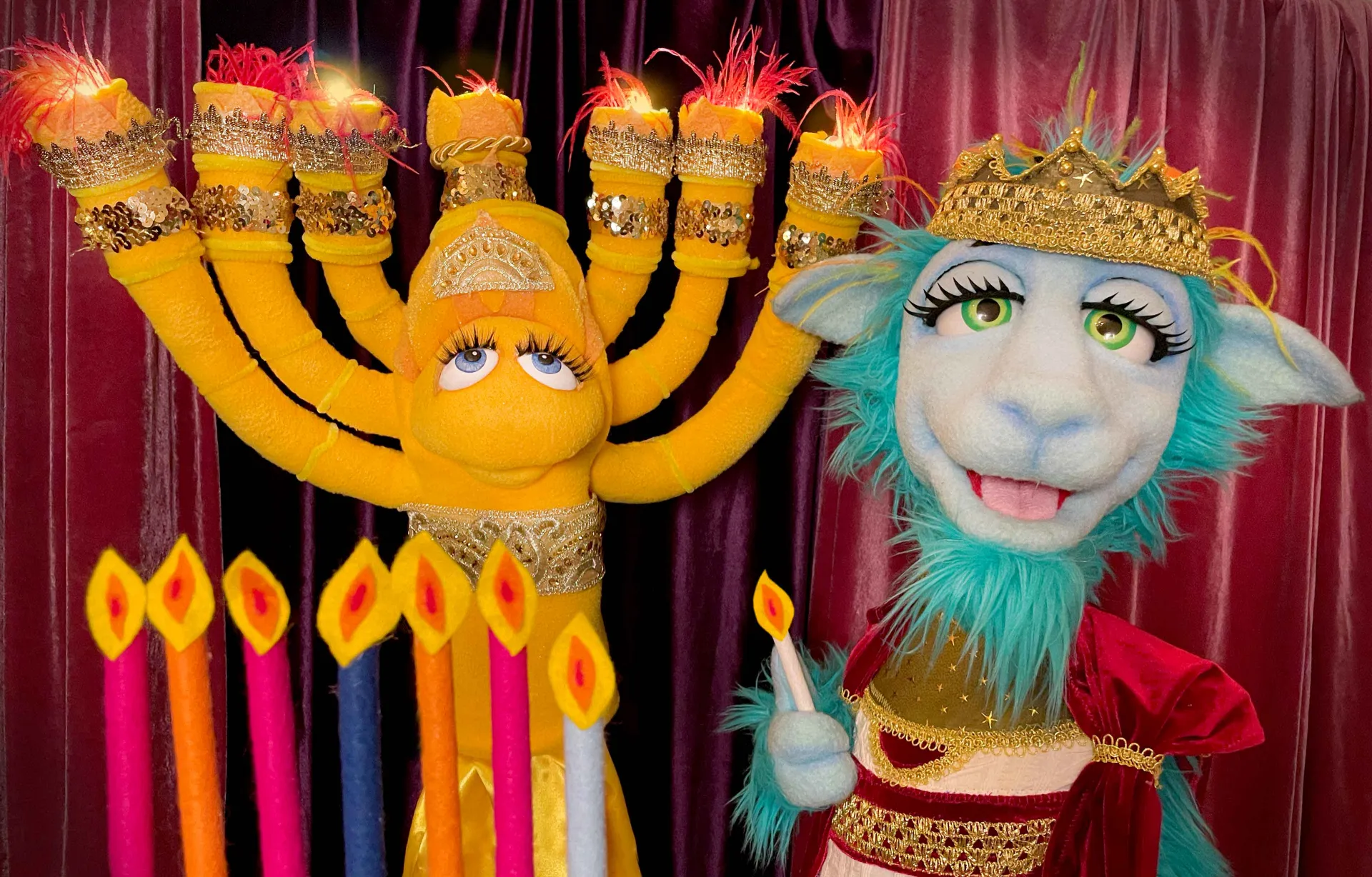 Zwei Handpuppen, eine in Form einer Chanukkia und eine als Schaf, stehen vor einem roten Bühnenvorhang mit acht Kerzen.