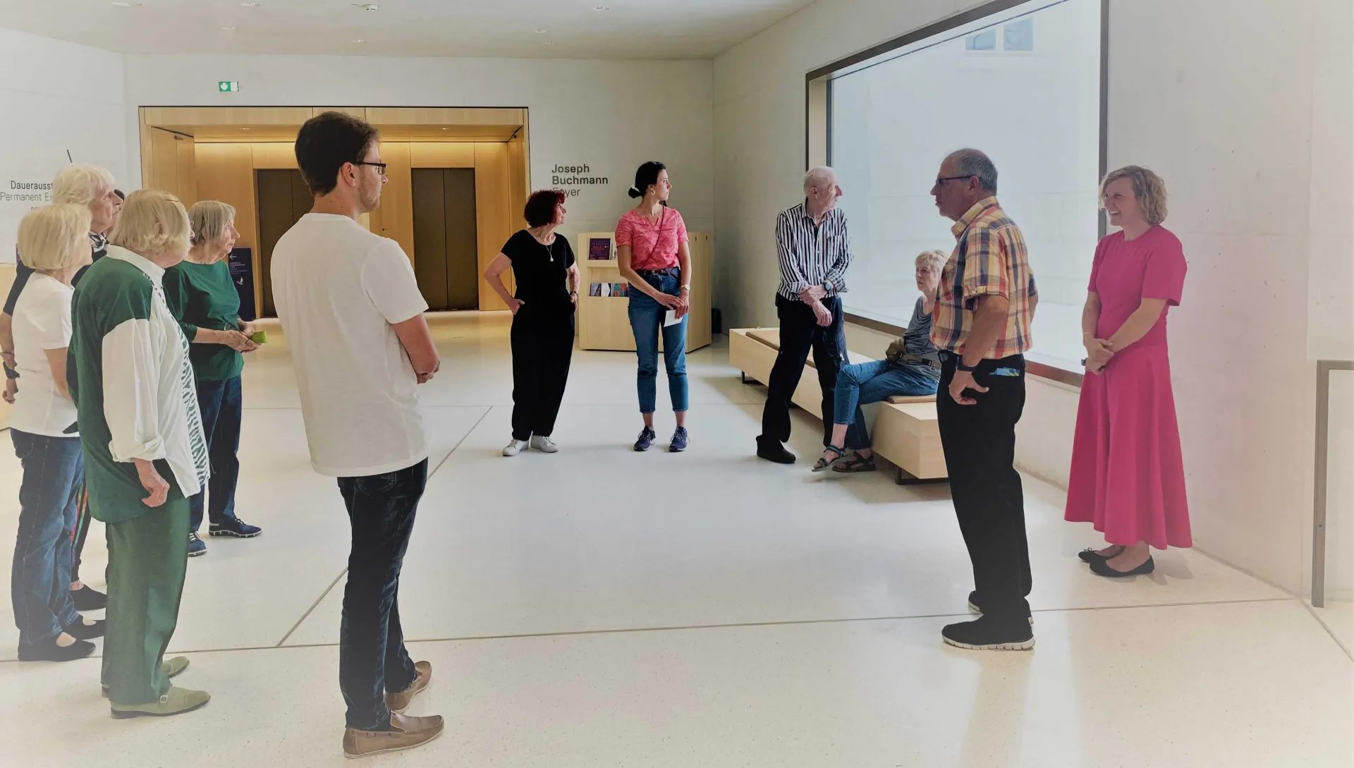 A group of people is standing in a white room with a large window. Most of the people are looking out of the window.