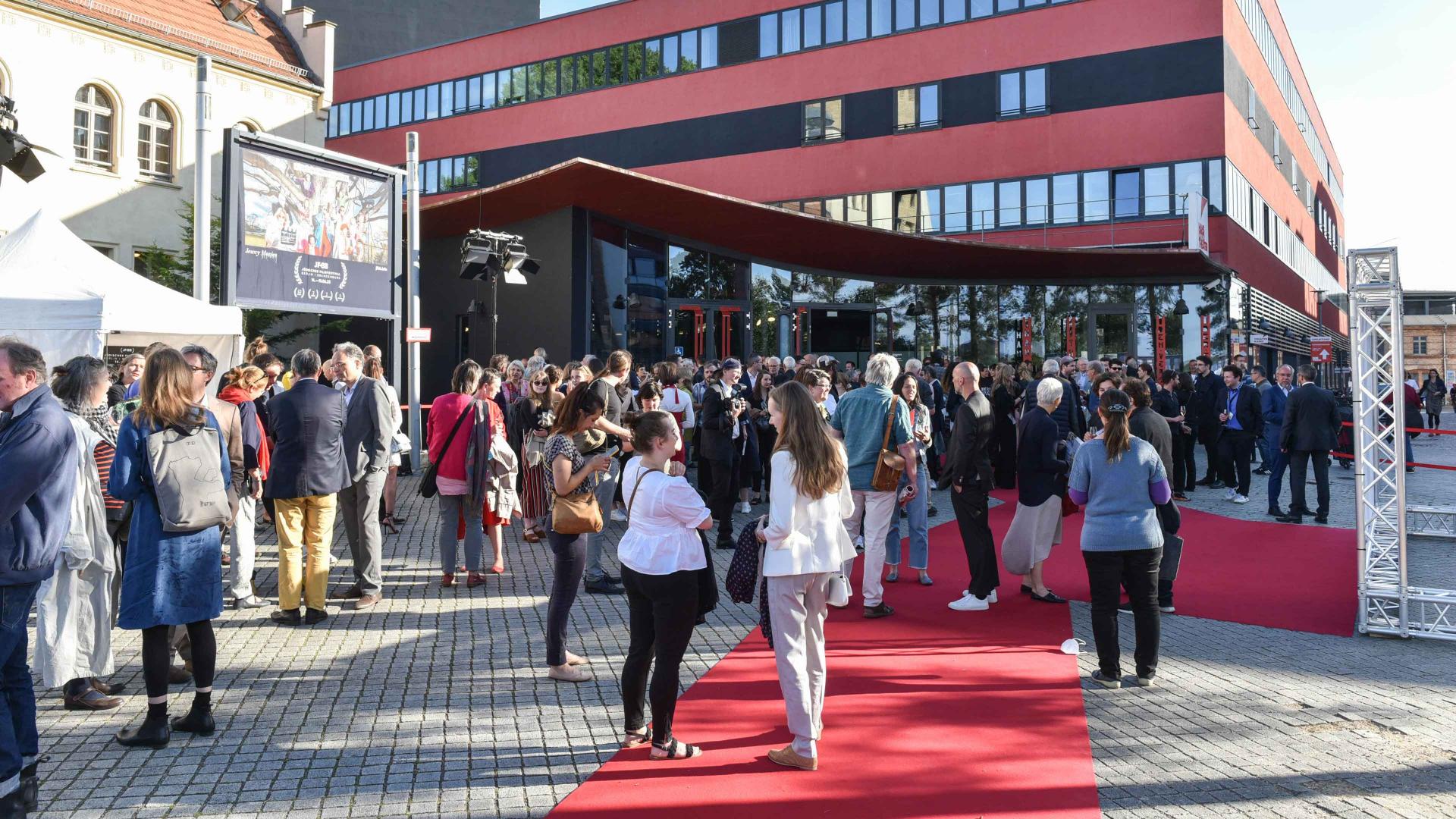 Red new building with people standing in front of it on a red carpet.