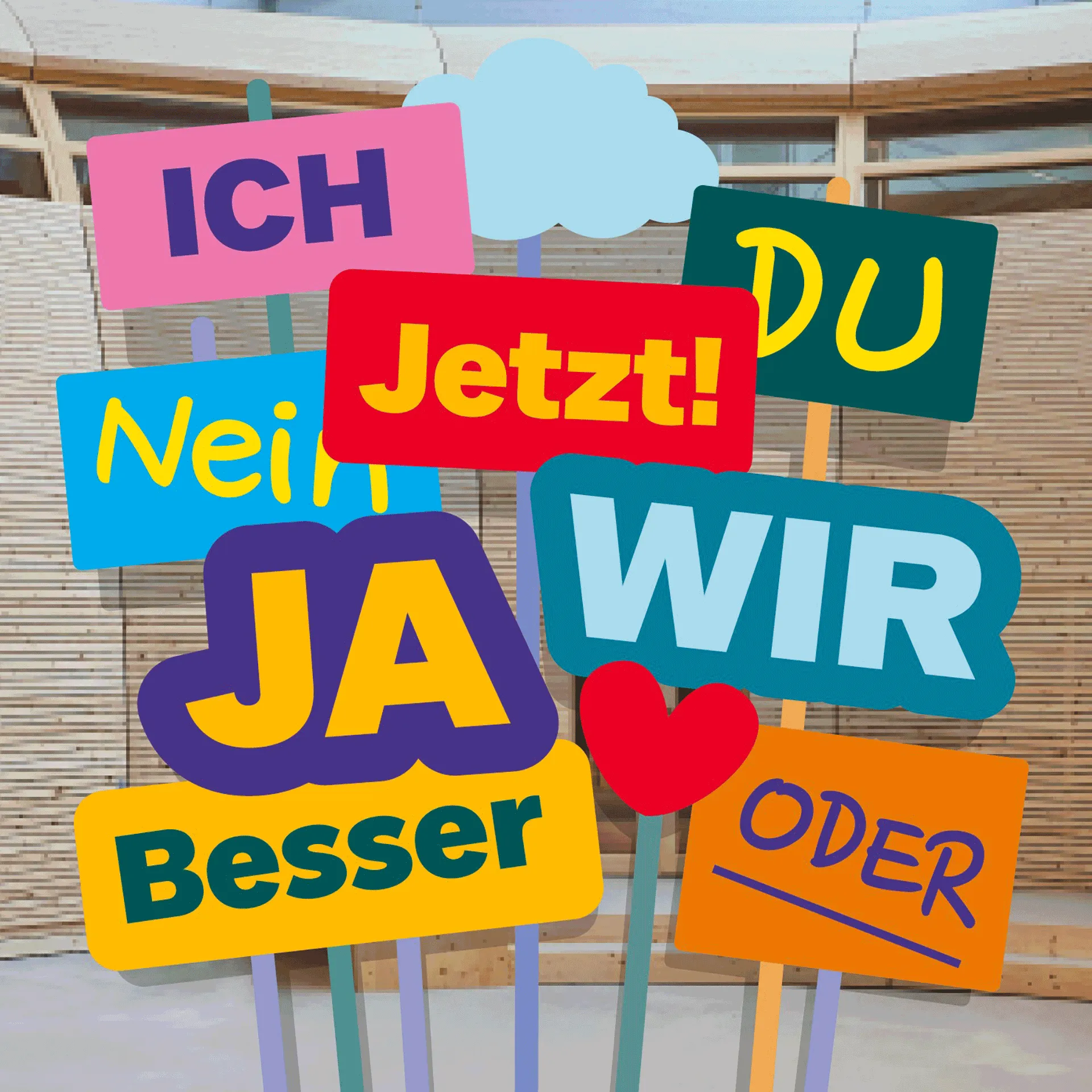 Bunte Grafik vor Holzarchitektur im ANOHA: stilisierte Demo-Plakate mit den Worten Ich, Du, Jetzt, Ja, Nein, Wir, Besser, Oder.