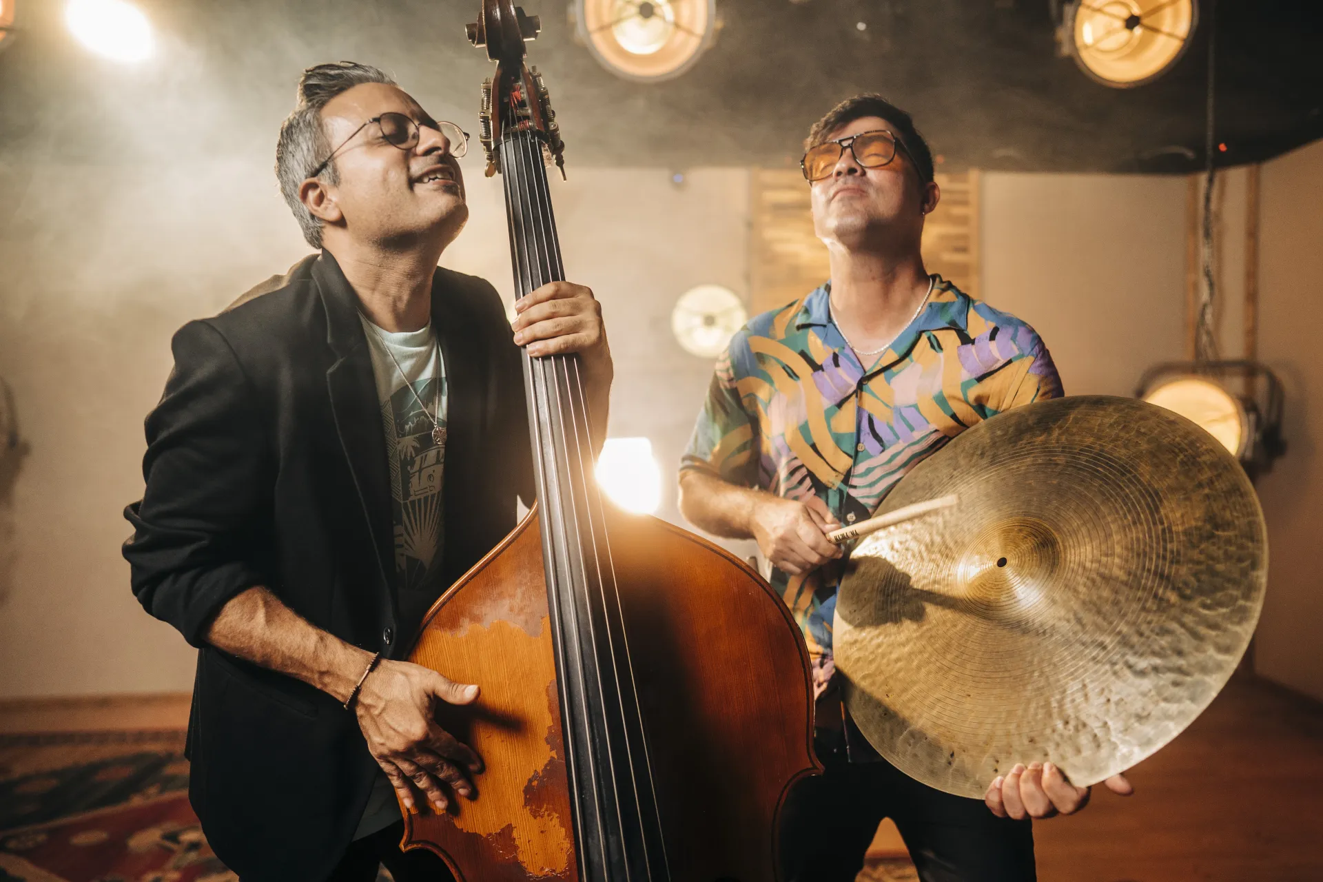 One man is playing bass, while the other is holding a cymbal in his hand. Both have their eyes closed. Spotlights are visible in the background.