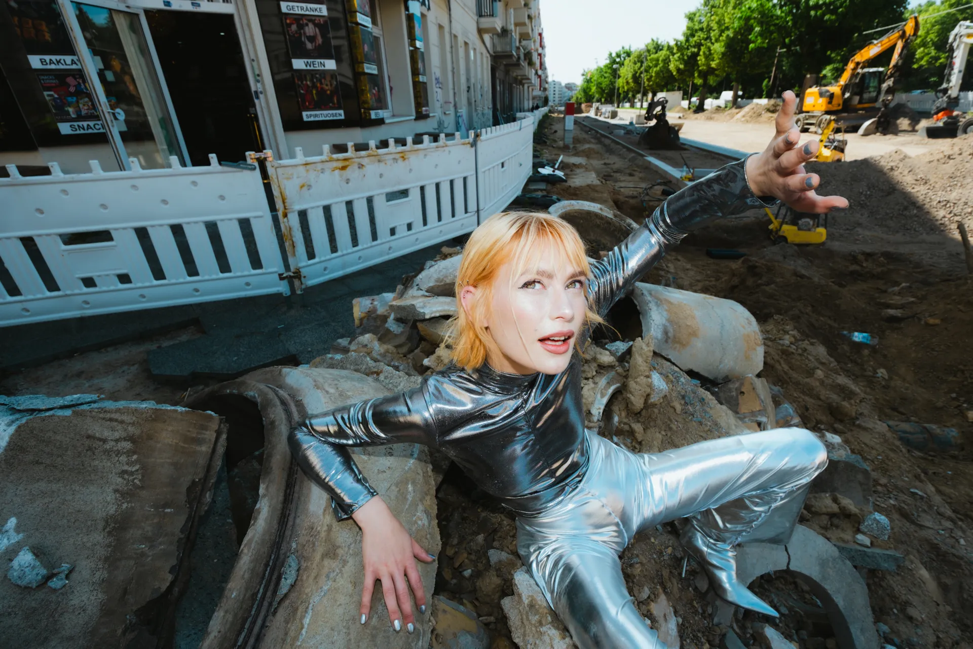 Frau in silberner Kleidung sitzt auf einer Baustelle, st&uuml;tzt sich auf einem Stein ab und h&auml;lt den anderen Arm nach oben, als w&uuml;rde sie geblendet.