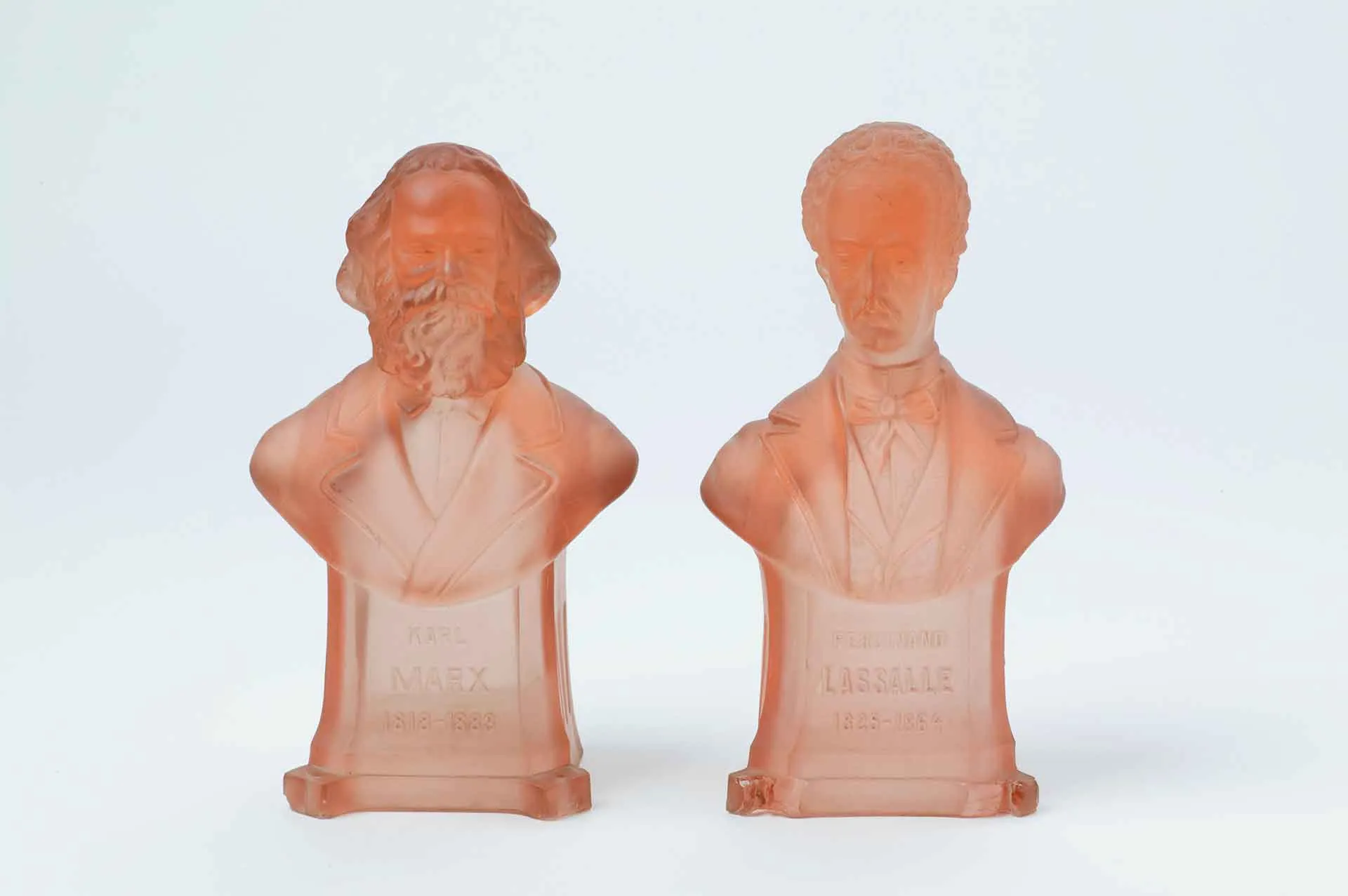 Two busts made of red, transparent glass. The one on the left depicts Karl Marx, the one on the right depicts Ferdinand Lassalle. 