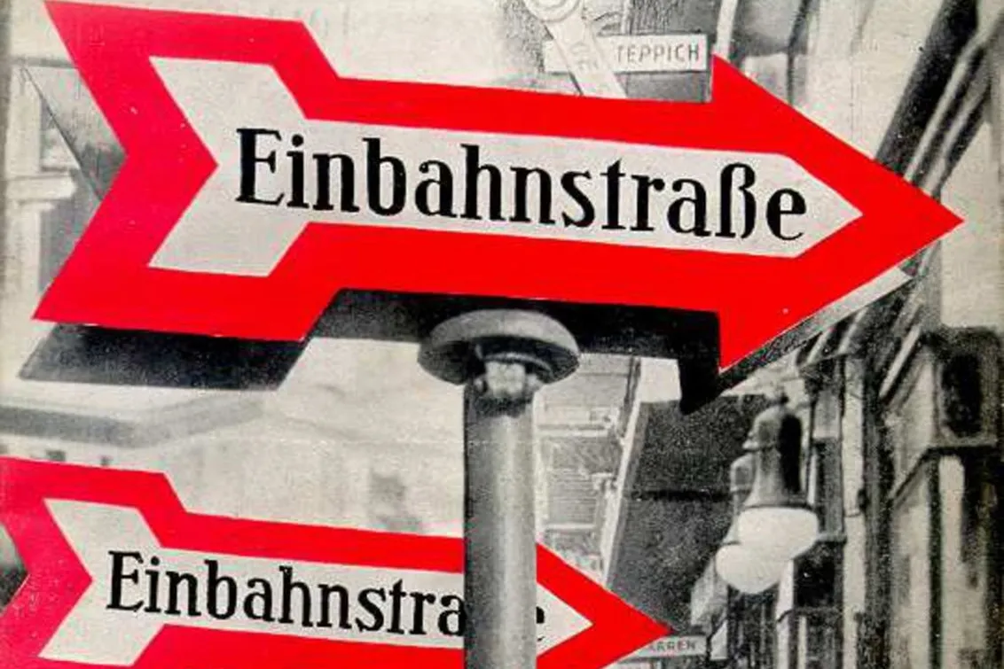 Book cover of One-Way Street by Walter Benjamin: A black-and-white photograph of a street; in the foreground, several traffic signs with a red arrow and the inscription One-Way Street.