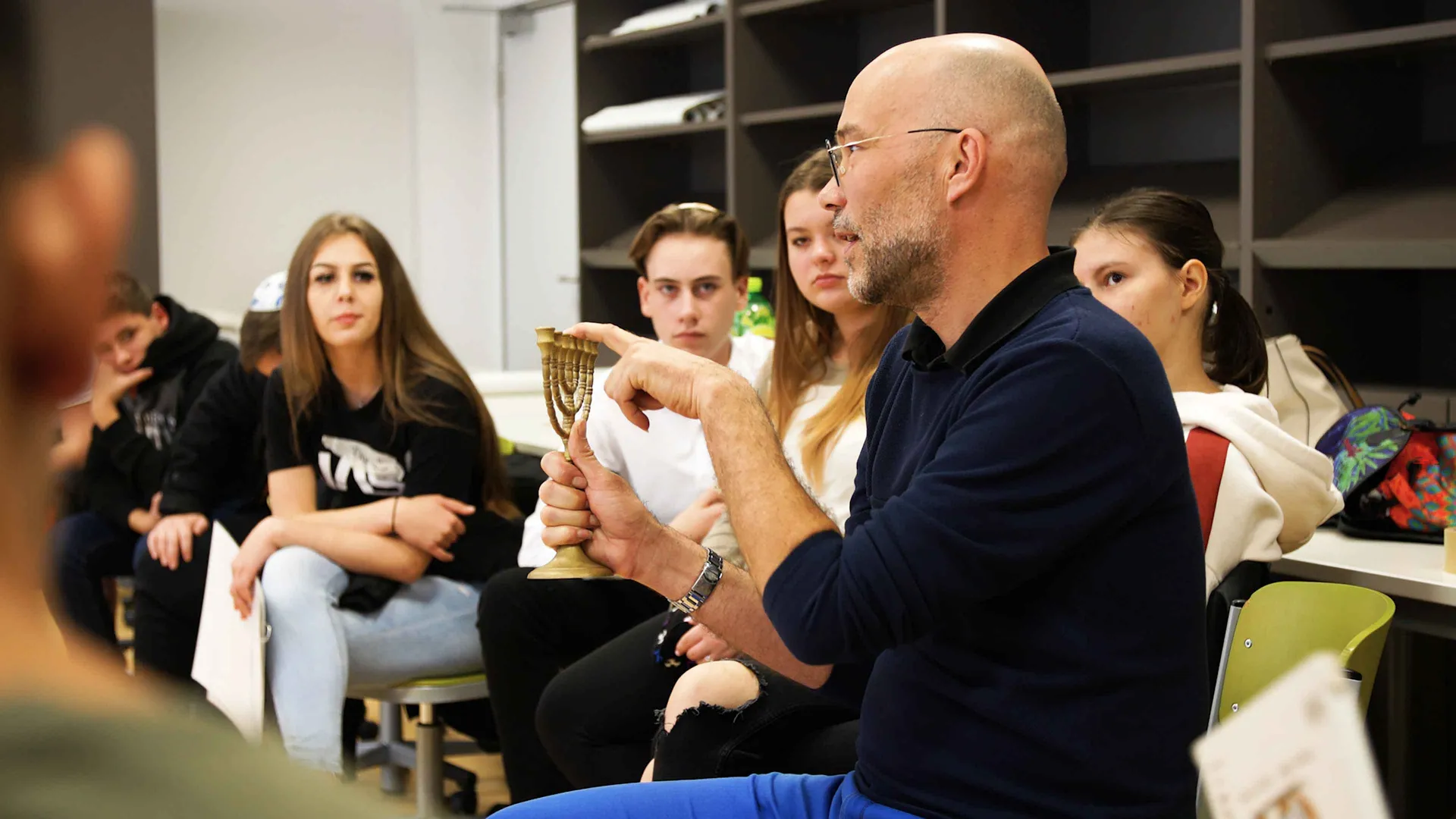 Ein Mann mit Brille sitzt zwischen Schüler*innen in einem Stuhlkreis und hält erklärend eine Menora, einen Kerzenleuchter, in der Hand.