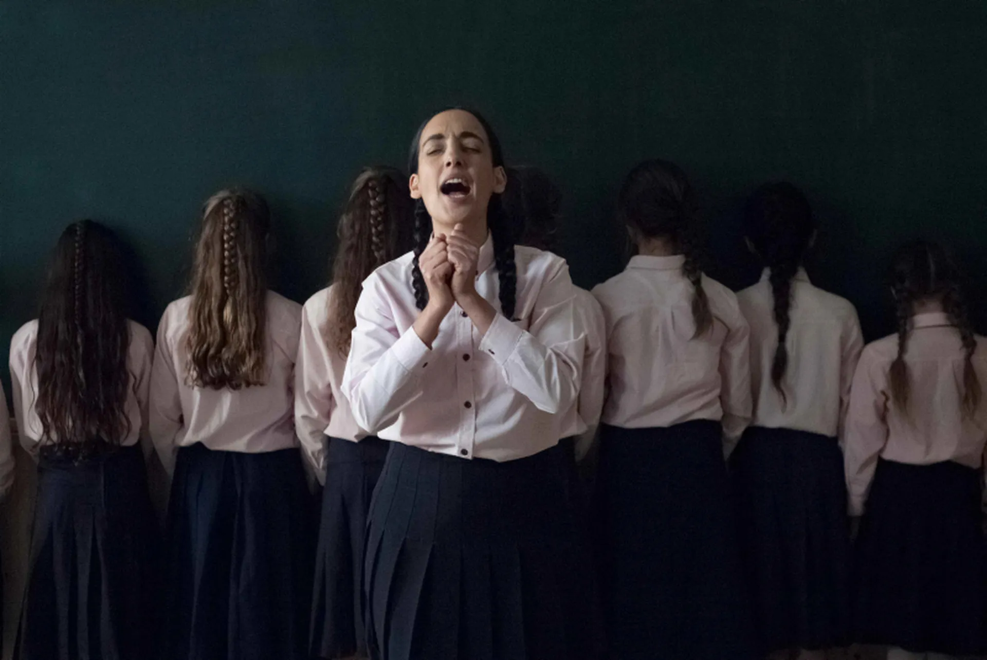Mehrere junge Frauen stehen in einer Reihe mit dem R&uuml;cken zur betrachtenden Person. Eine junge Frau steht vorne, leicht vor der Reihe, frontal zur Kamera. Sie hat die Augen geschlossen, den Mund ge&ouml;ffnet und h&auml;lt ihre H&auml;nde senkrecht vor sich, leicht zu F&auml;usten geballt, sodass sie sich ber&uuml;hren.