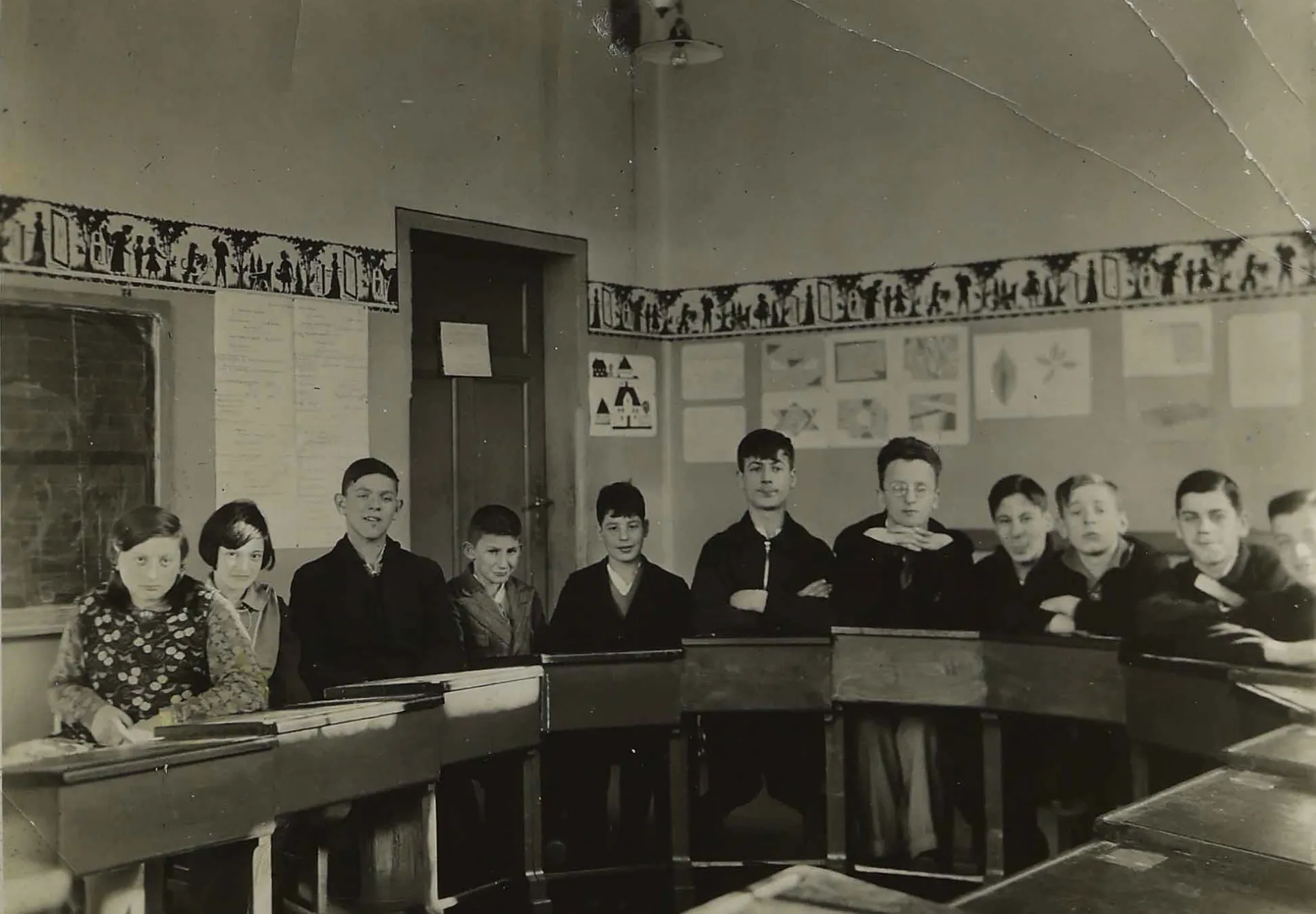 Das Bild zeigt einen Schulklassenraum mit elf Sch&uuml;lerinnen und Sch&uuml;lern, die in einem Halbkreis angeordnet an ihren Pulten sitzen.