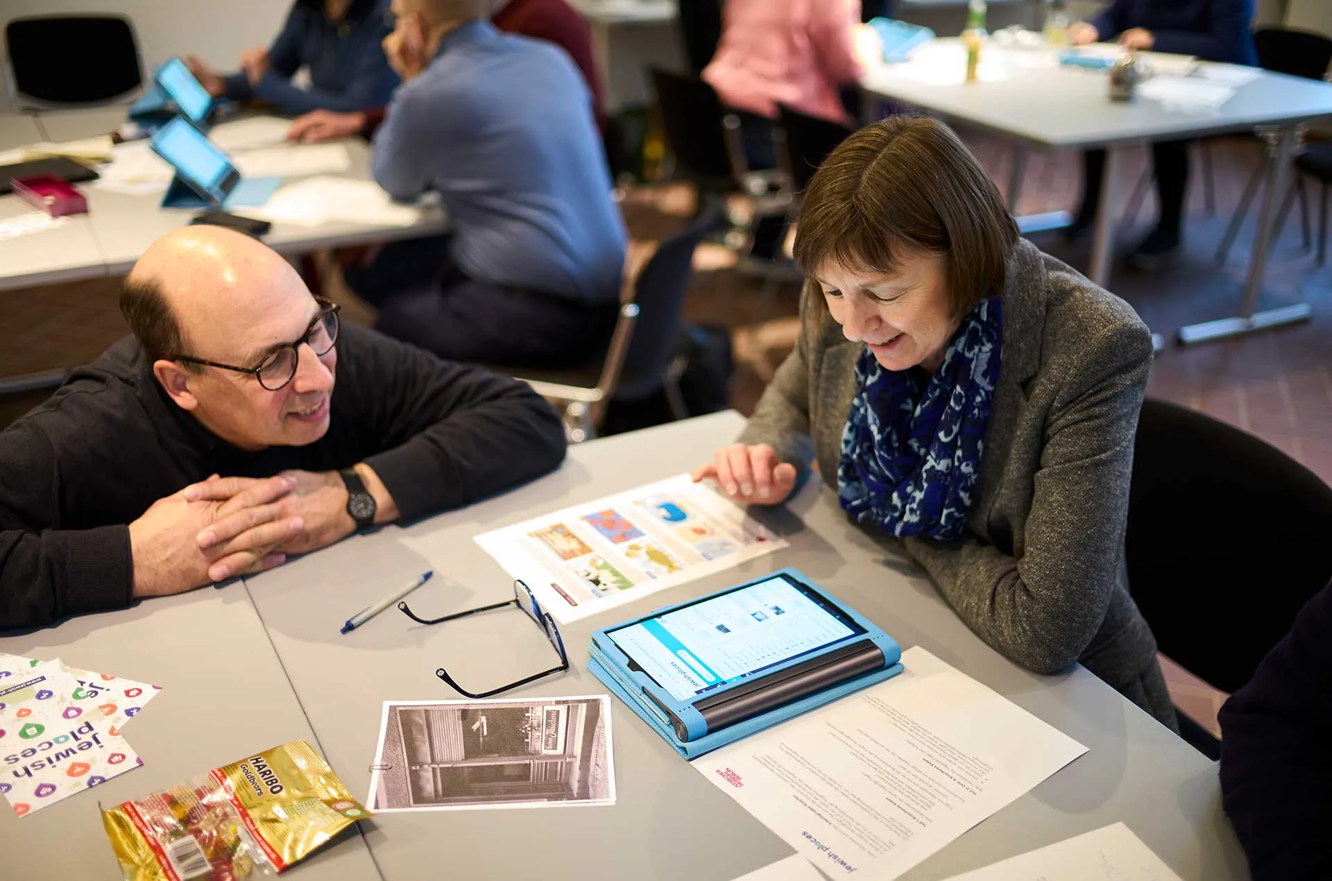 Zwei Personen sitzen an einem Tisch in einem Workshop- oder Seminarraum und betrachten gemeinsam Unterlagen sowie ein Tablet. Auf dem Tisch liegen ausgedruckte Materialien, eine Brille und weitere Arbeitsunterlagen. Im Hintergrund arbeiten weitere Teilnehmende an Tischen.