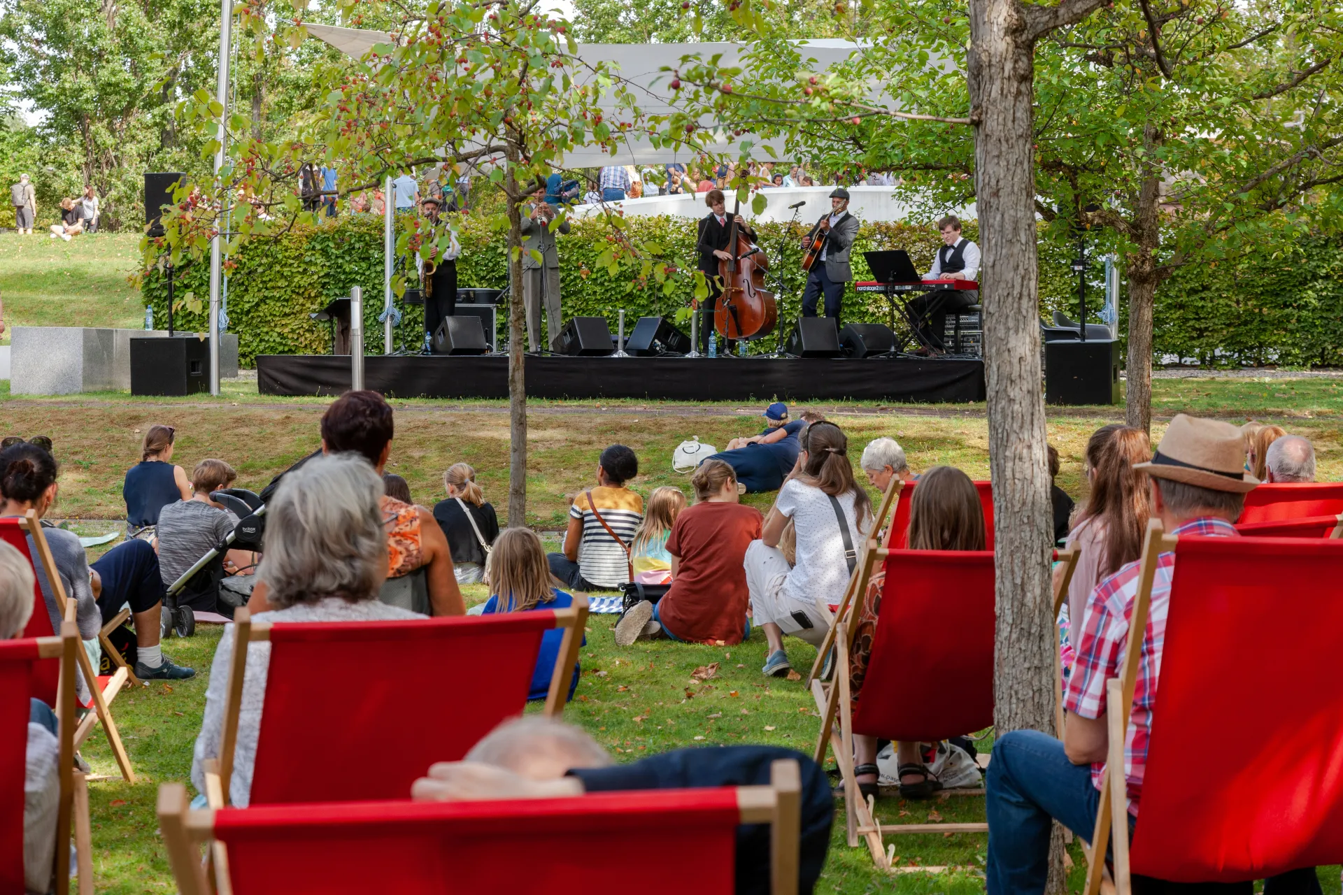 The audience lies on lounge chairs in a garden and watches a music band performing on a stage.