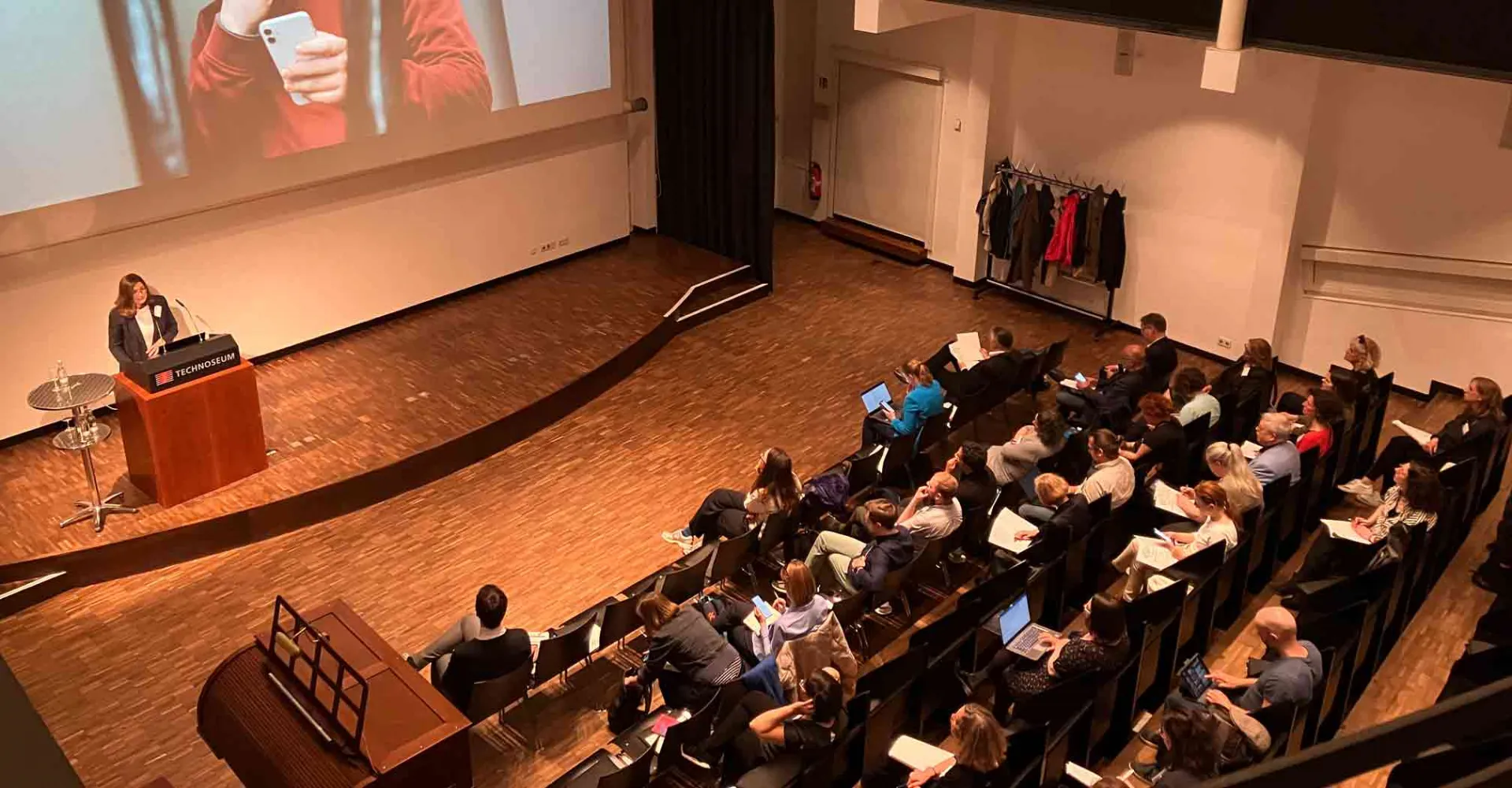 Vollbesetzter Hörsaal mit Rednerin am Pult und auf der Leinwand hinter ihr ein junger Mann mit Kopfhörern und Smartphone.