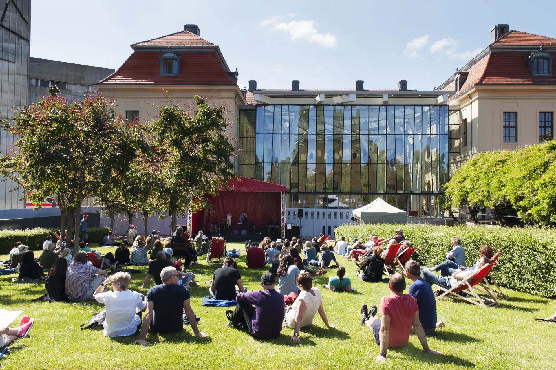 Several people are sitting on the lawn in the museum garden, looking toward the stage.
