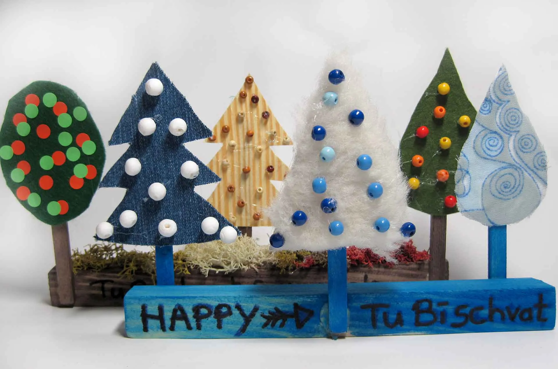 Das Bild zeigt sechs selbst gebastelte, geschm&uuml;ckte B&auml;umchen aus Holz und Pappe, die an kleine Holzleisten geklebt sind. Auf den Leisten steht: "Happy TuBischvat". 