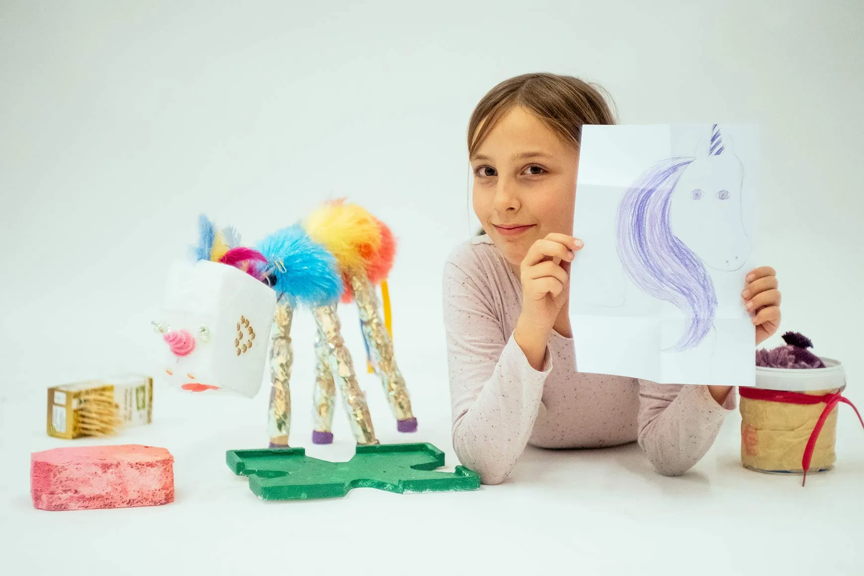 A child next to a handcrafted unicorn holds a drawing of a unicorn in the camera.