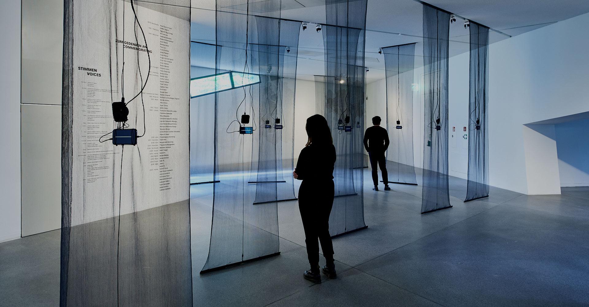 View of the exhibition with visitors, from the ceiling hang black transparent fabric panels, to each of which is attached a smartphone with sound waves on the display