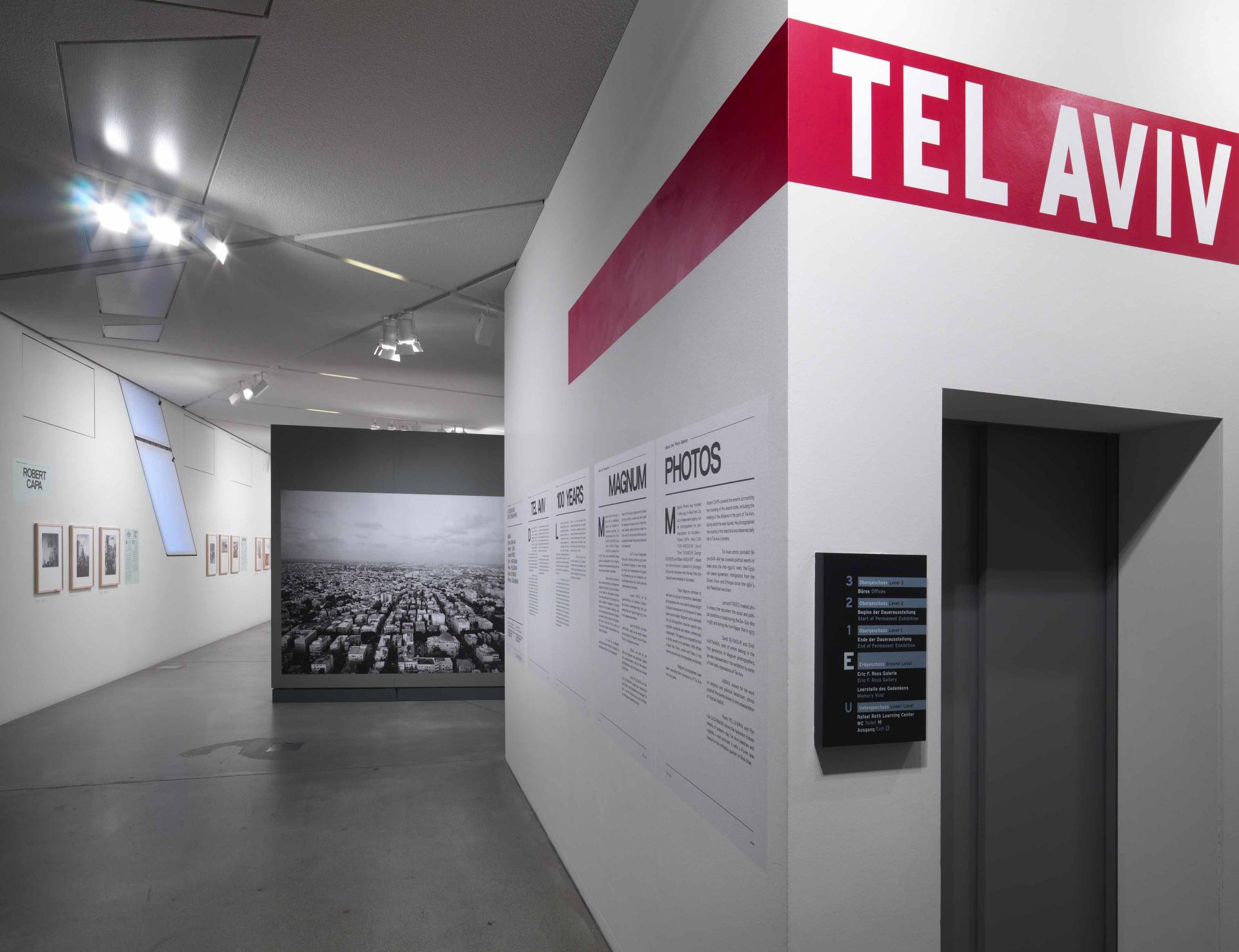 View into the entrance area of the exhibition: On the right a wall with lettering, in the background a large black-and-white photo showing an aerial view of a city