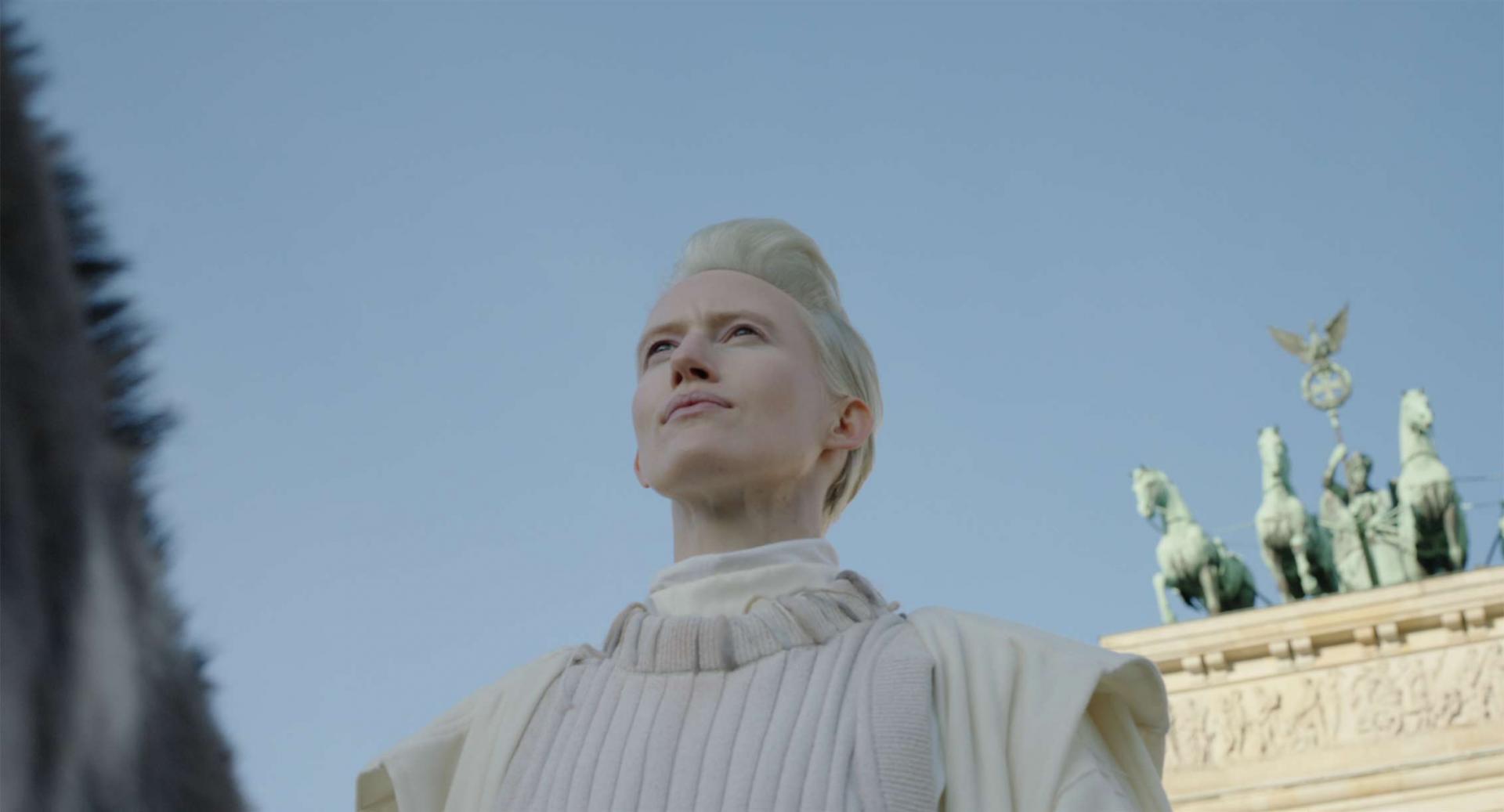 Worm's-eye view of the chest and face of a woman dressed in white with platinum blonde, short hair on a donkey, whose neck, cut at the left edge of the picture, is just visible. Background: Berlin Brandenburg Gate and steel blue sky