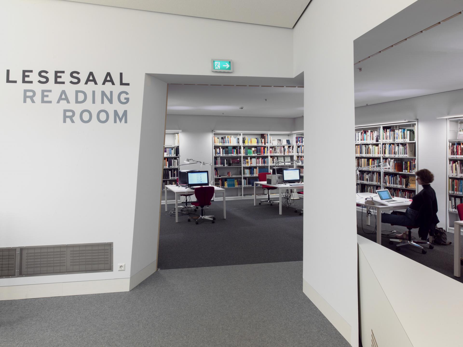 Passage from the reading room to the work area, a woman sits at one of the computers on the tables