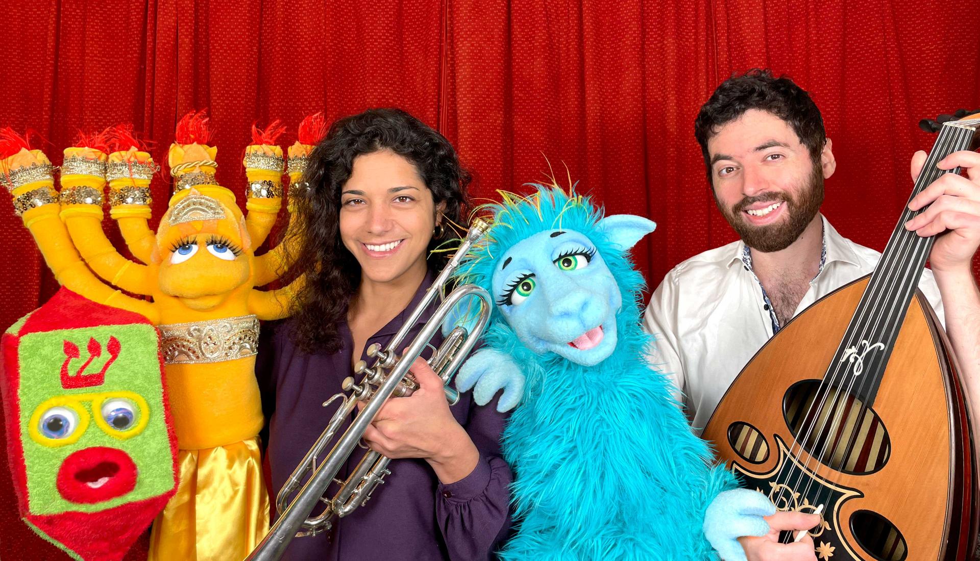Two people holding instruments are surrounded by three plush creatures in the form of a menorah, a dreidel and an animal.