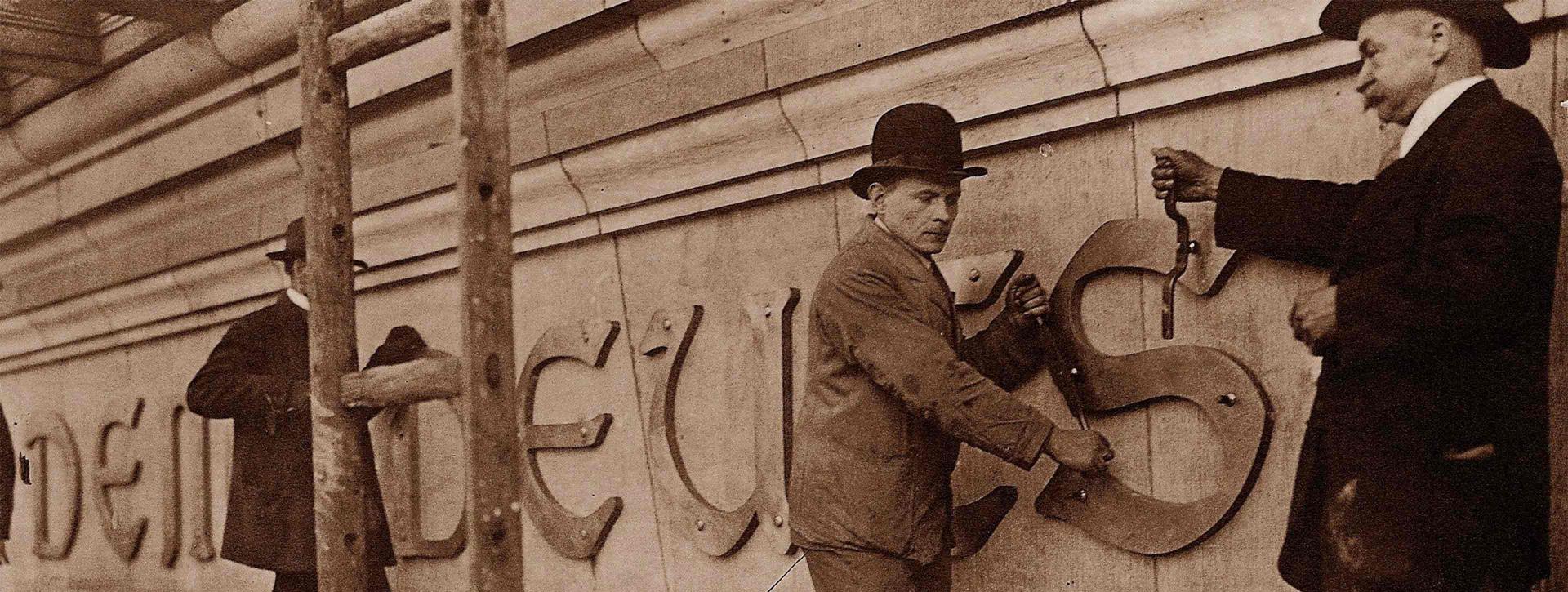 Handwerker mit Werkzeug bringen einen Schriftzug an einer Fassade an.