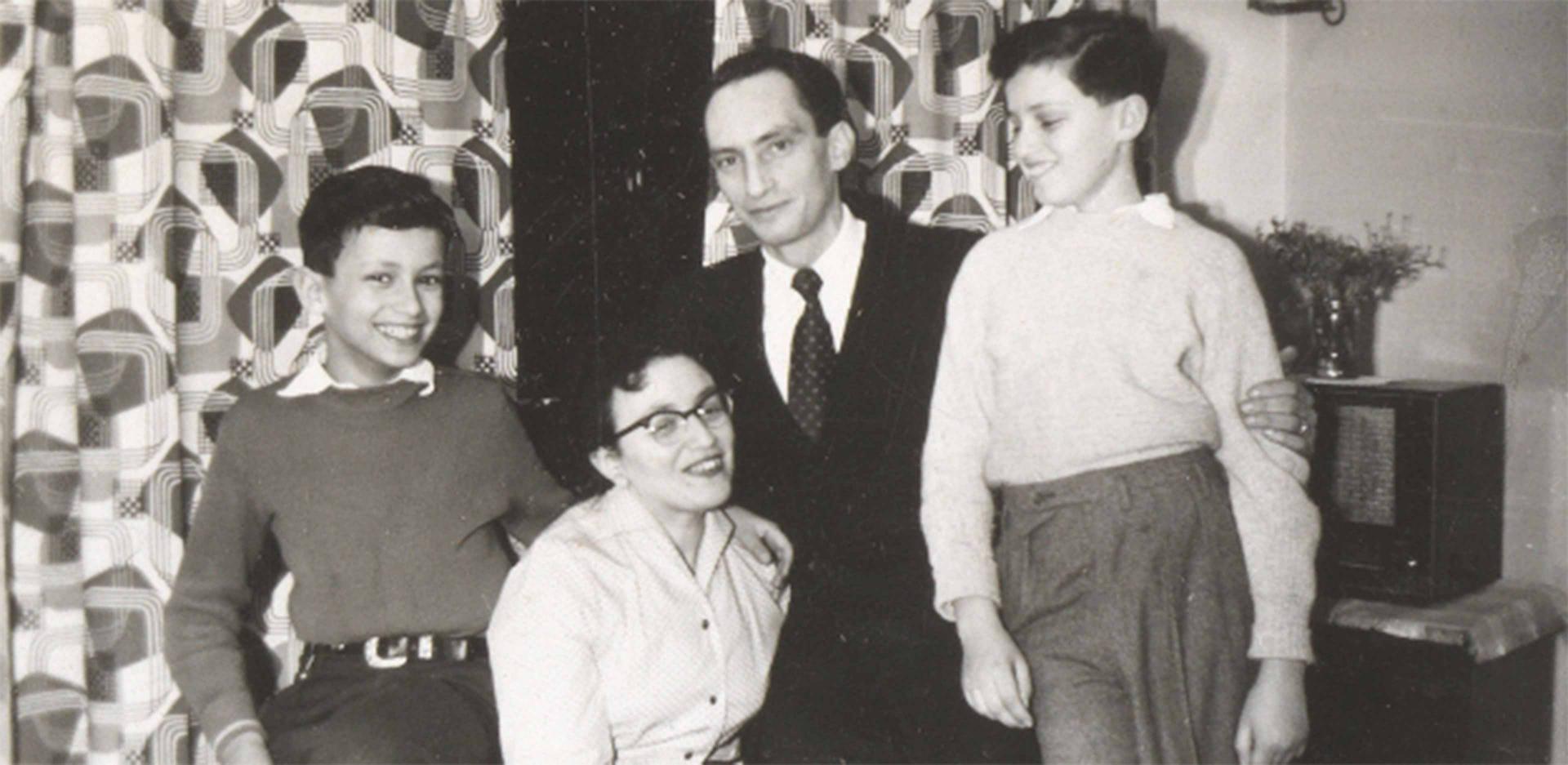 Die Aufnahme zeigt zwei Erwachsene und zwei Kindern in einer Wohnung mit gemusterten Vorhängen. Die beiden Jungs stehen rechts und links neben den Erwachsenen, in der Mitte sitzt eine Frau mit Brille, rechts neben ihr befindet sich ein Mann im Anzug.